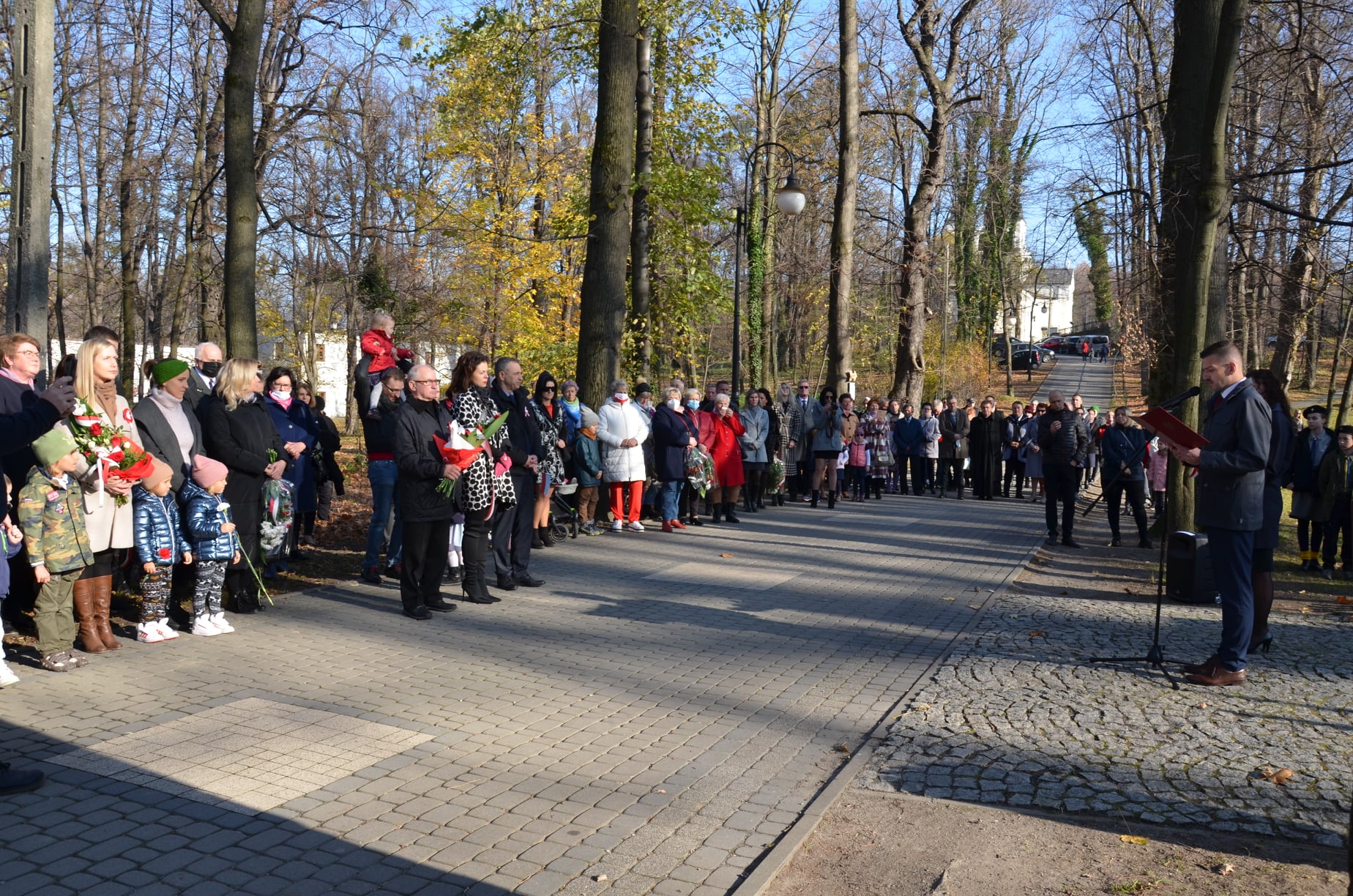 Grupa ludzi stojąca w parku i słuchająca przemówienia. Część z nich trzyma w rękach wiązanki kwiatowe.