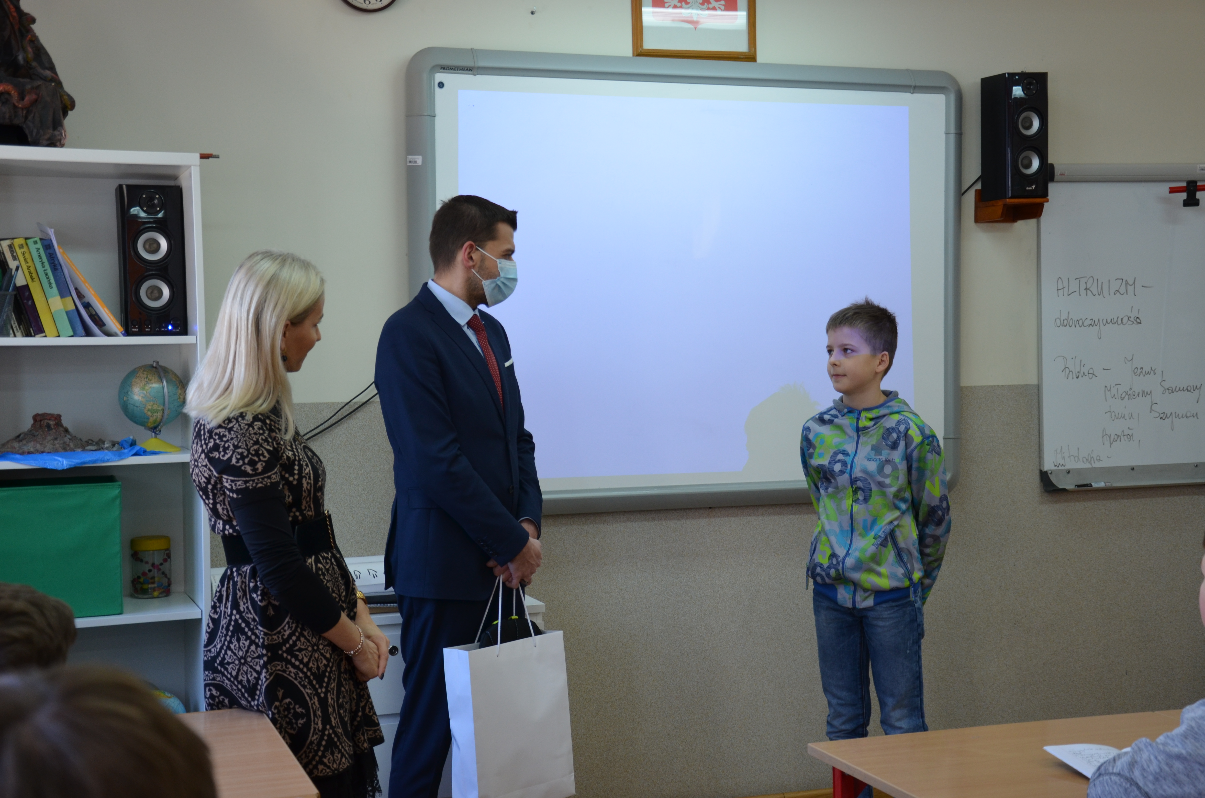 Trzy osoby stojące w sali szkolnej. Dwie z lewej, kobieta i mężczyzna, wręczają upominek nastolatkowi.