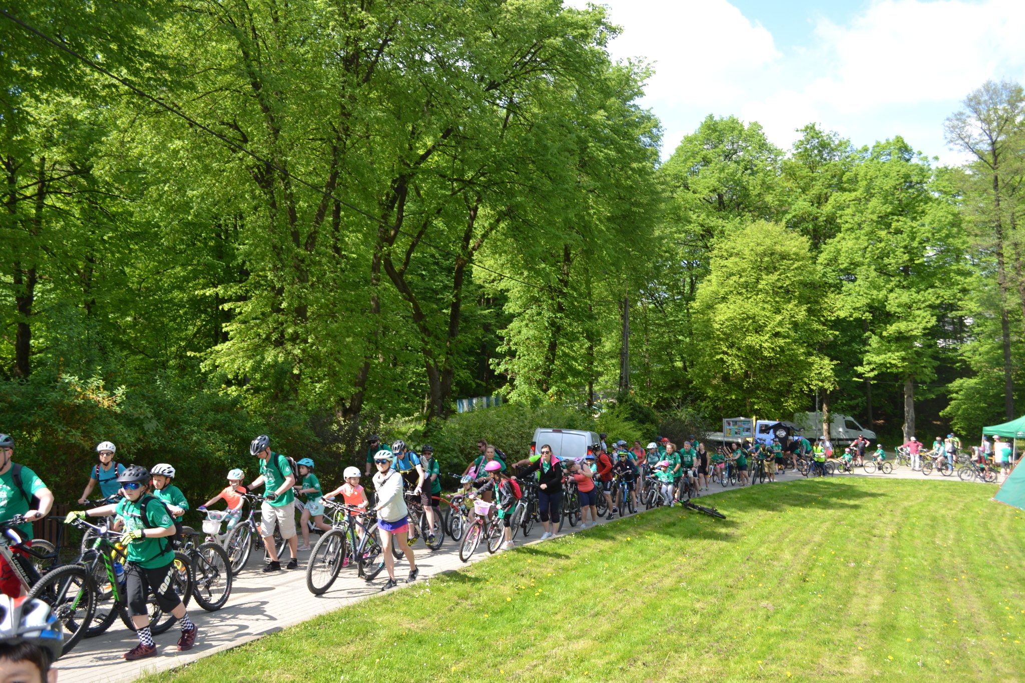Duża grupa rowerzystów jadących drogą wśród zieleni. Z tyłu szpaler drzew.