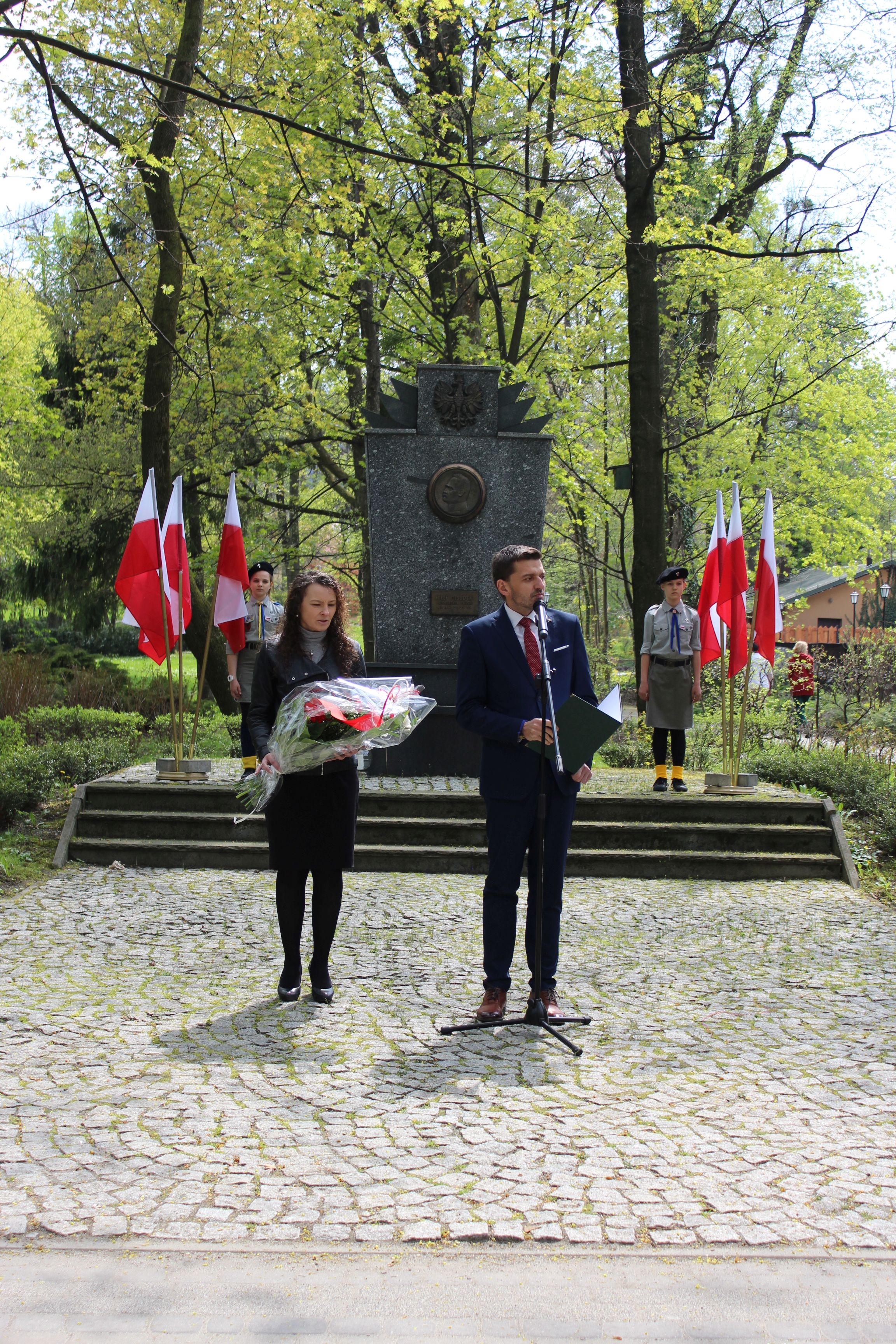 Wójt i Zastępca Wójta stojący przed pomnikiem, z tyłu dwoje harcerzy. Obok nich flagi Polski.