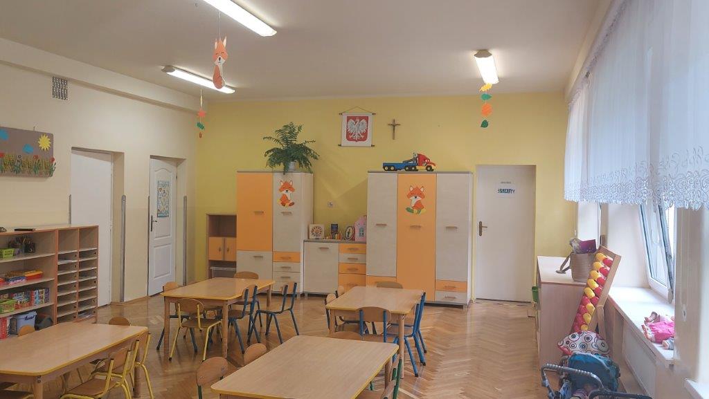 Sala przedszkolna, drewniany parkiet, na ścianie wisi godło Polski, pod ścianą szafki, na środku ławki