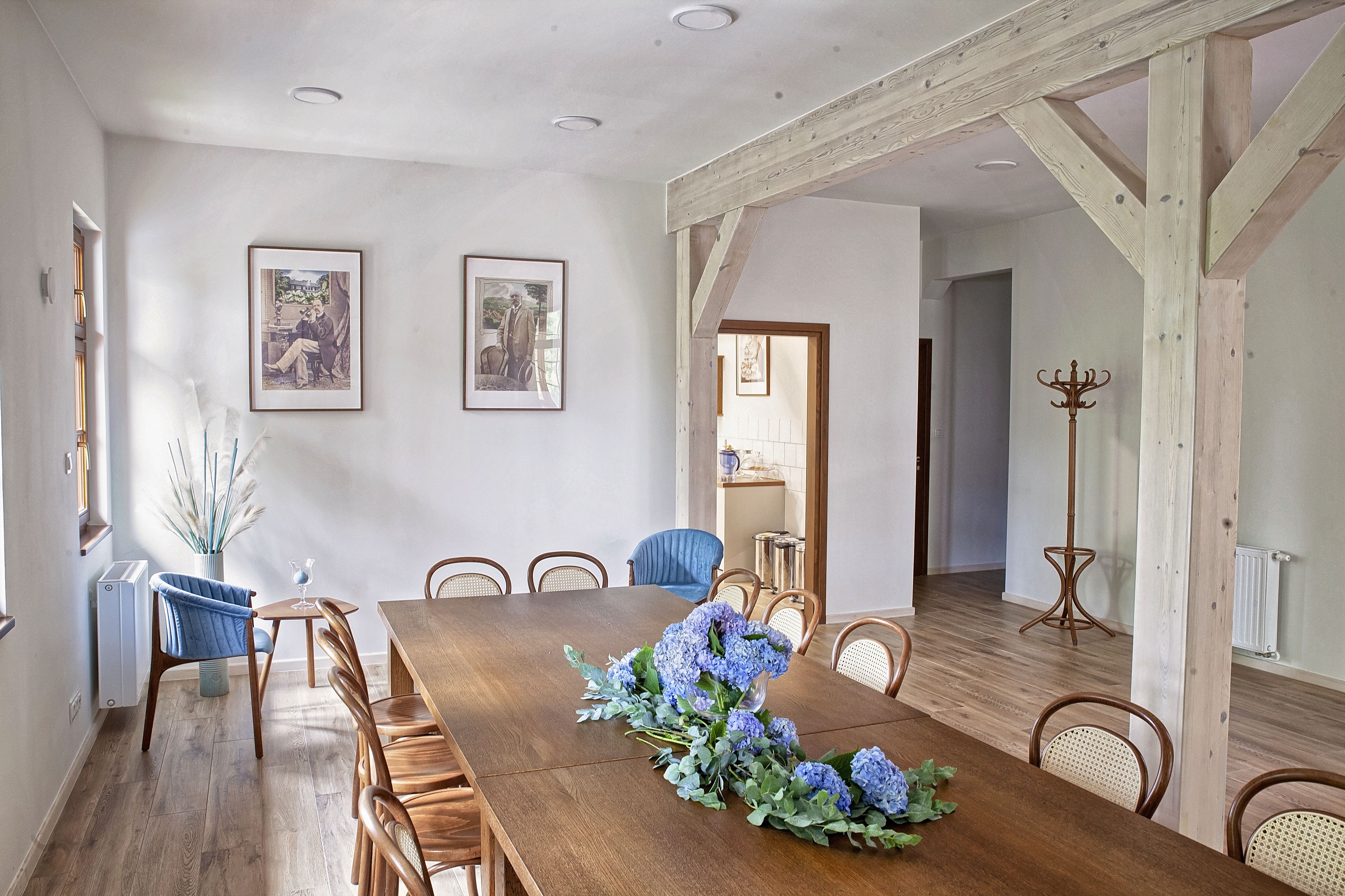 długi drewniany stół z krzesłami, na stole kwiaty, w tle widok na wiszące na ścianie obrazy