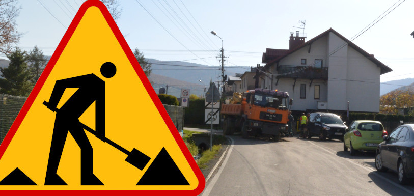 Po lewej znak drogowy przedstawiający człowieka z łopatą, po prawej droga, po której porusza się samochód ciężarowy z koparką.