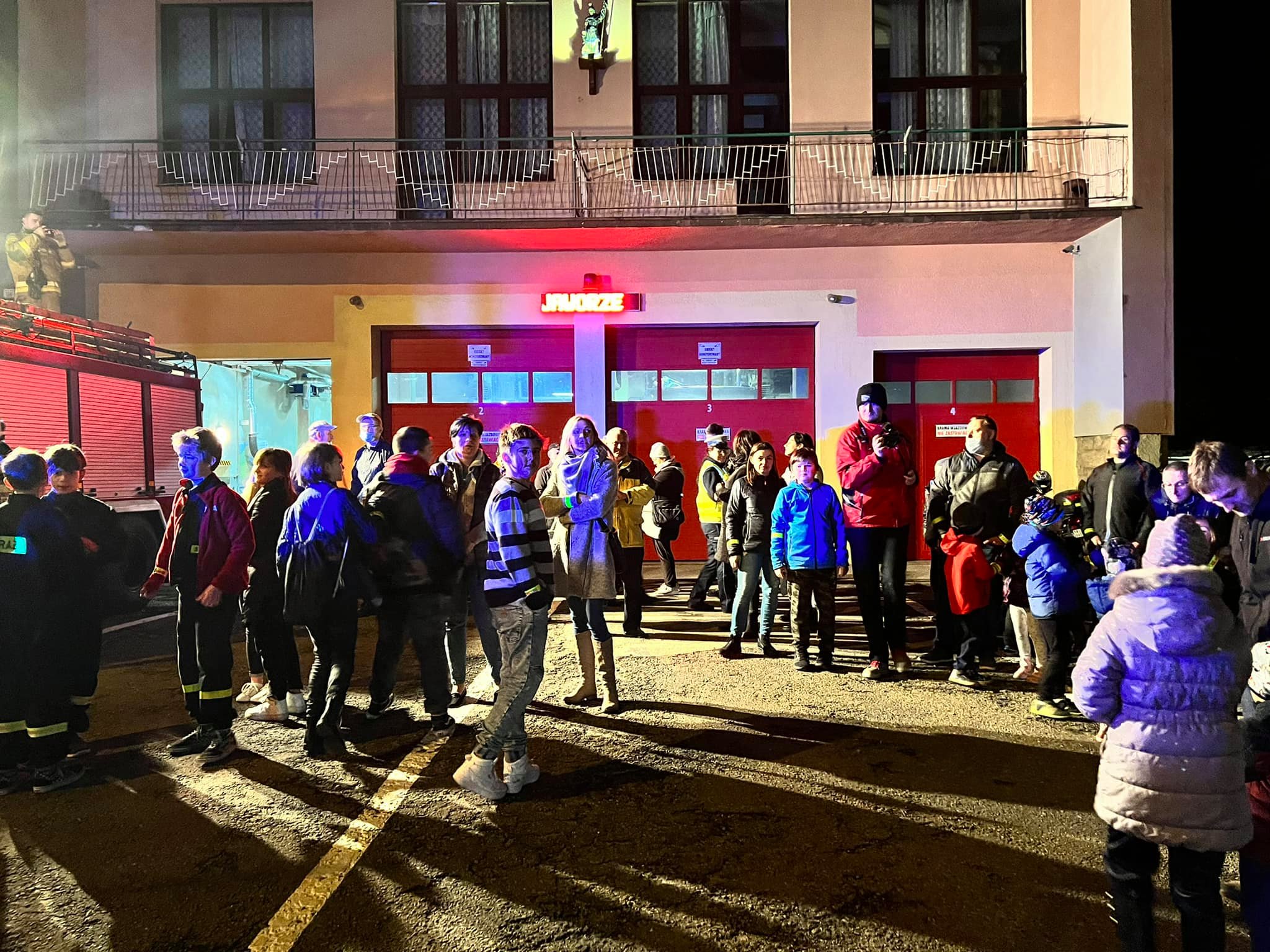Grupa ludzi stojąca przed budynkiem straży pożarnej - zdjęcie nocne.