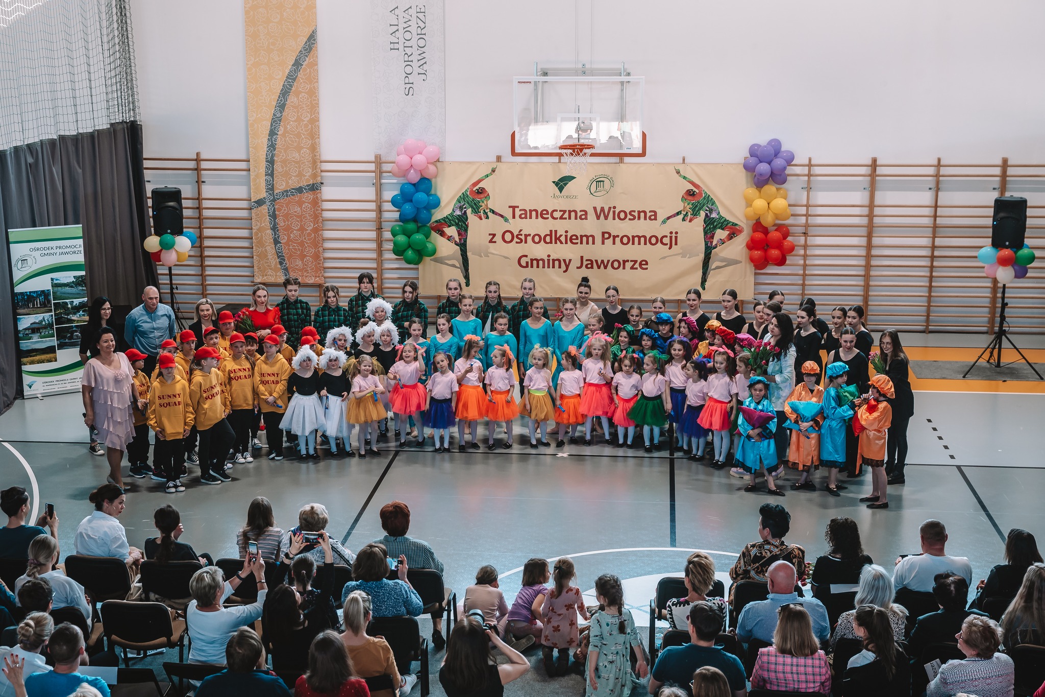 Grupa kilkudziesięcioro dzieci ubranych w różnorakie stroje na hali sportowej.
