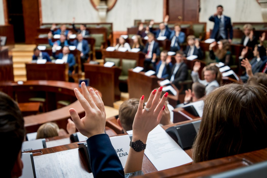 Dwie podniesione ręce w akcie głosowania, w tle sala posiedzeń