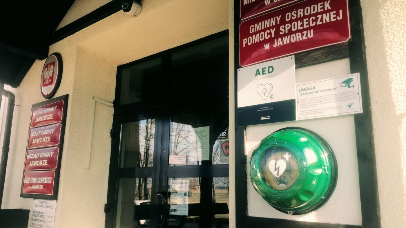 Wejście do budynku Urzędu Gminy. Po prawej, pod tabliczkami informacyjnymi podwieszony okrągły defibrylator AED.