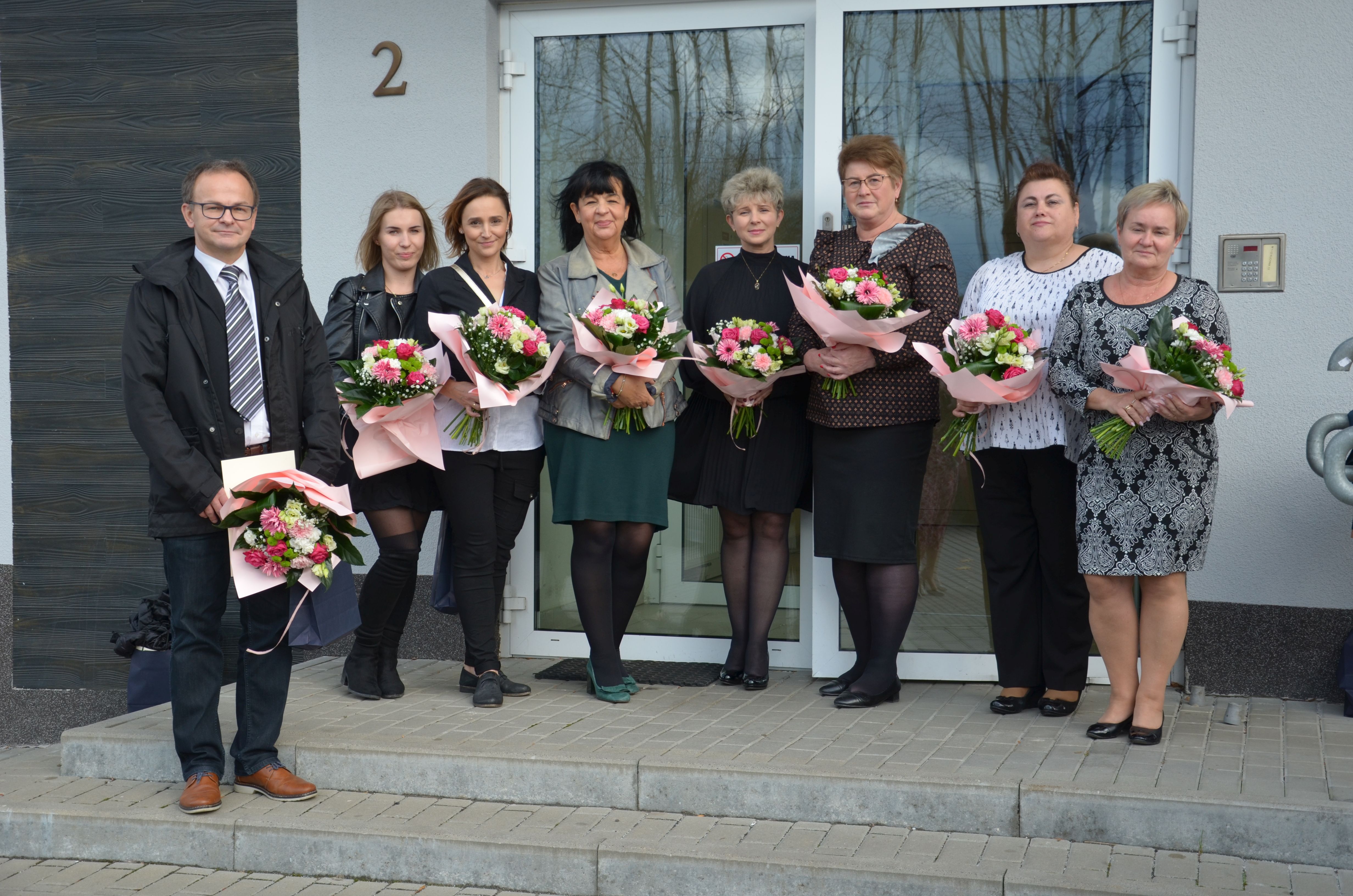Grupa 8 osób (mężczyzna i kobiety) stojąca przed wejściem do budynku. W ręce trzymają kwiaty.